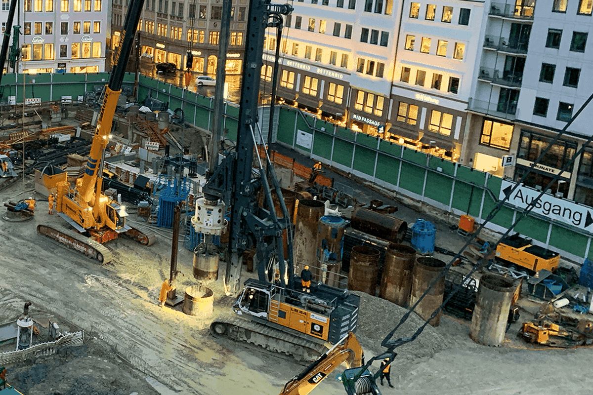 Eine Baustelle mit Maschinen und Arbeitern, umgeben von Gebäuden und einem Sicherheitszaun.