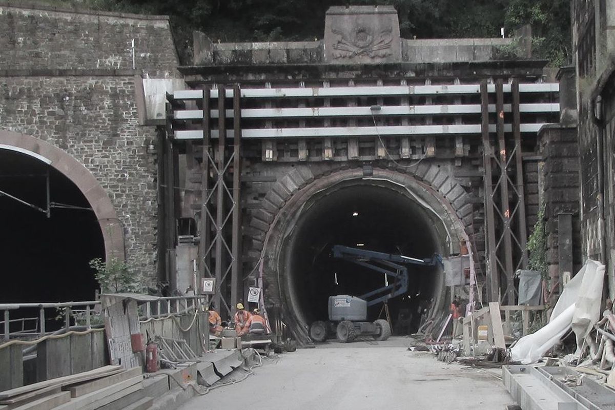 Blick auf zwei Tunnelöffnungen, eine Baustelle im Vordergrund mit Baugeräten und Arbeitern.