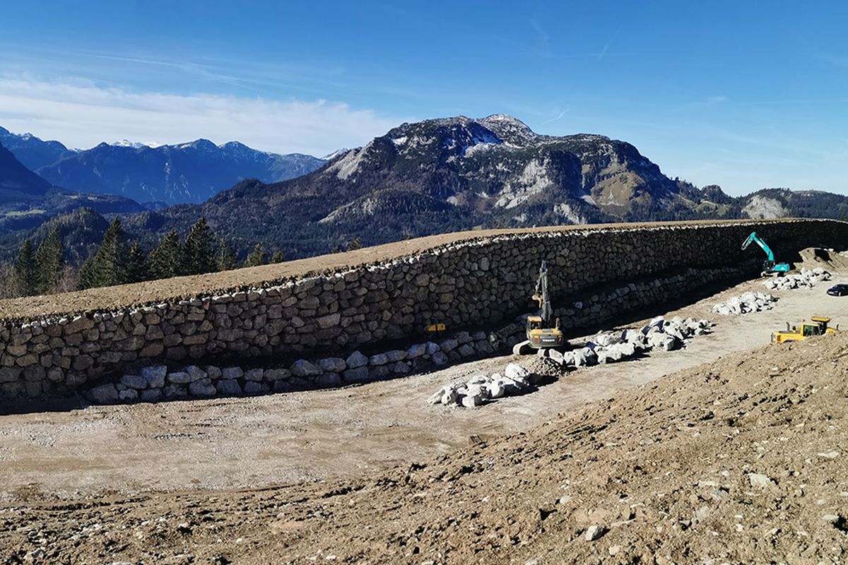 Panoramablick auf eine steinerne Mauer in den Bergen, umgeben von Bäumen, mit Baumaschinen und ohne menschliche Anwesenheit.