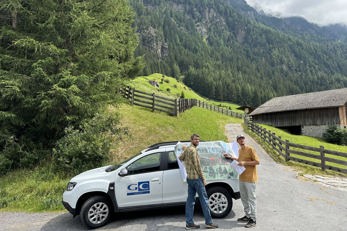 Zwei Personen betrachten eine Karte an einem Auto in einer ländlichen Umgebung mit Bergen und Wäldern.