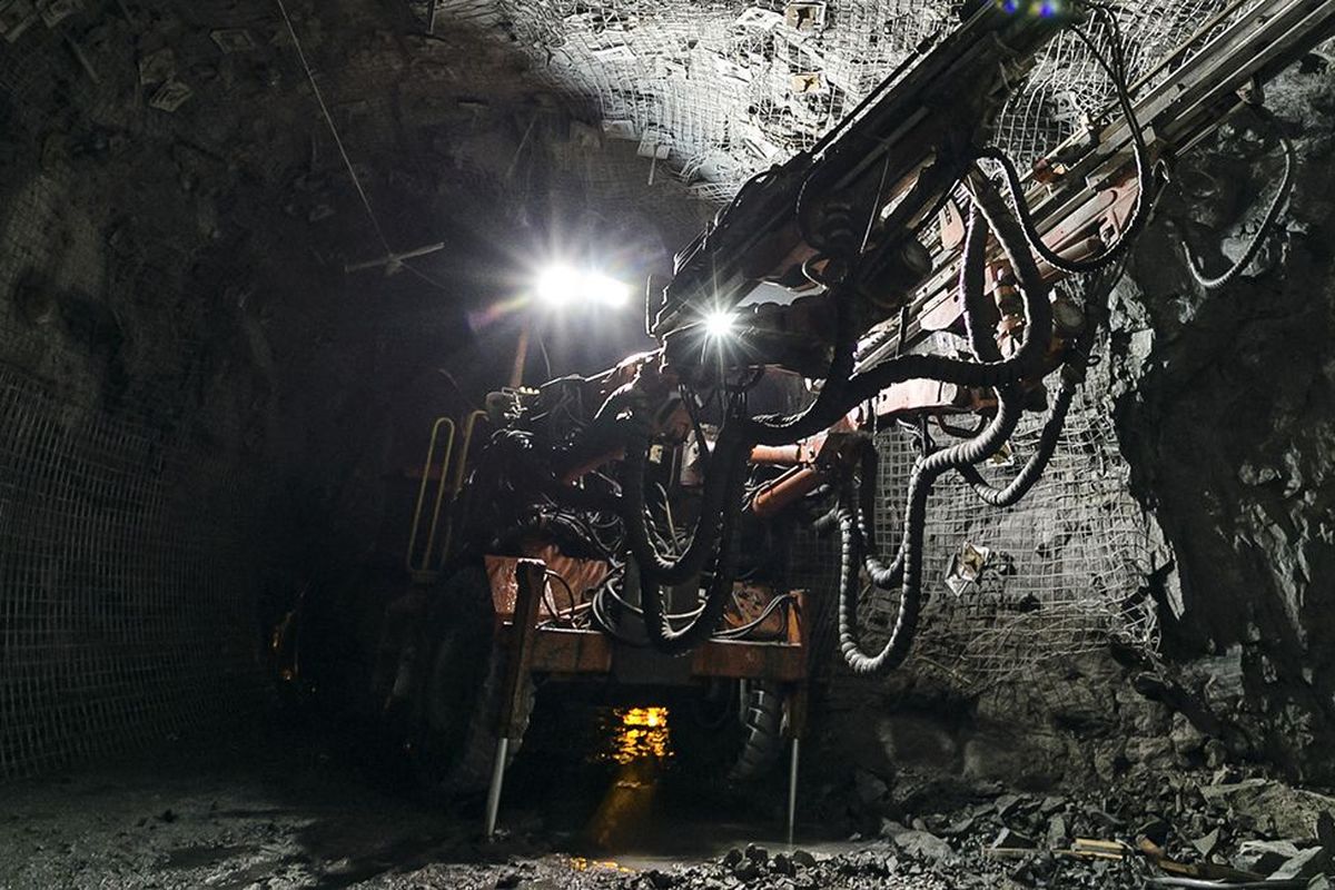 Ein Bergbaumaschinen in einem dunklen Tunnel, beleuchtet von Scheinwerfern, umgeben von felsigem Terrain.