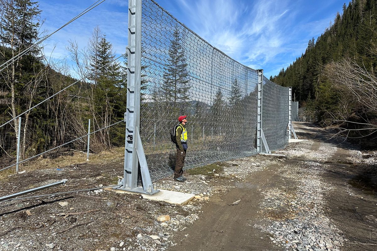 Eine Person steht an einem hohen Zaun aus Drahtgeflecht in einer ländlichen Umgebung mit Bäumen und einem blauen Himmel.