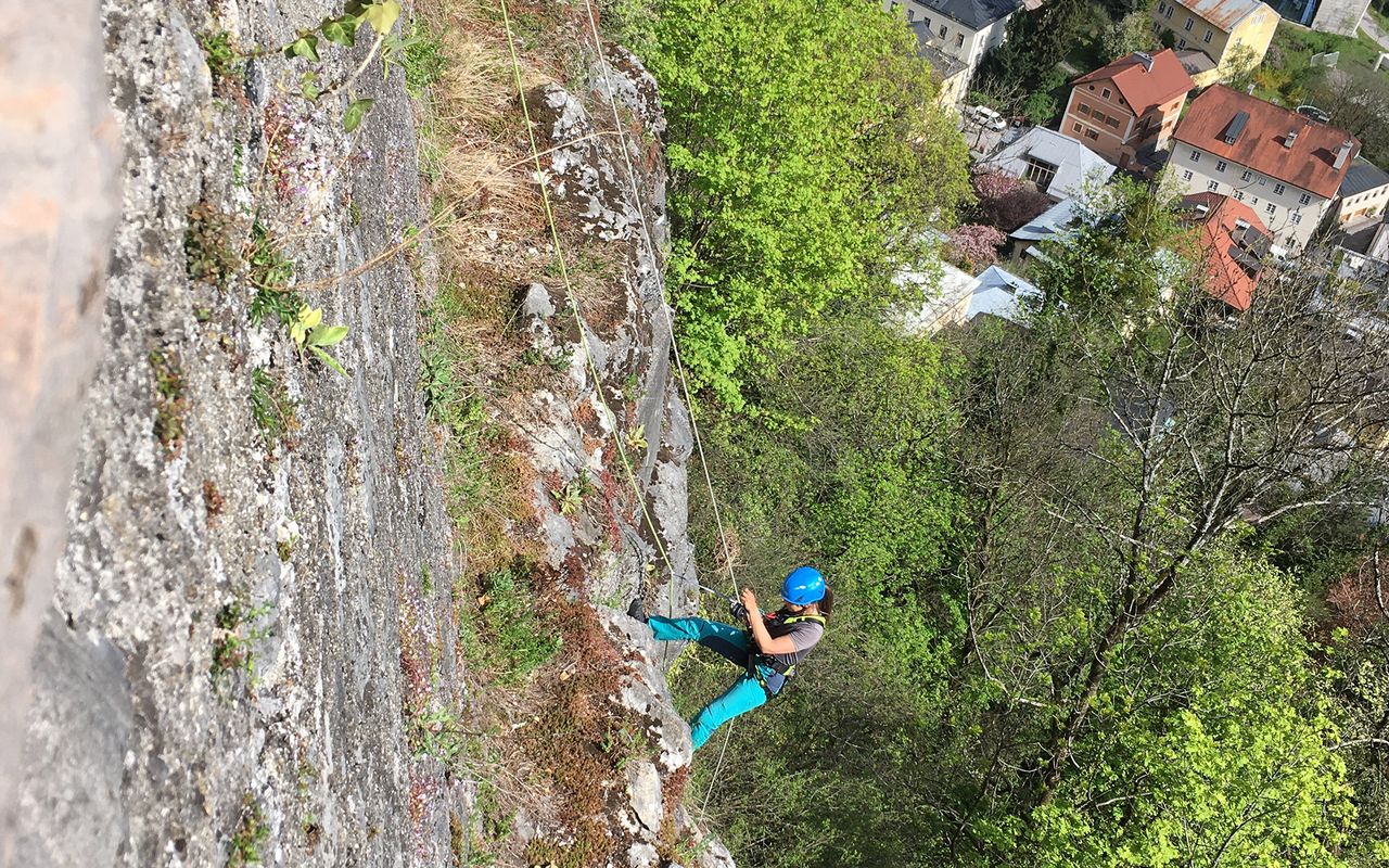 Eine Person klettert an einer steilen Felswand, umgeben von Bäumen und einer Stadt im Hintergrund.