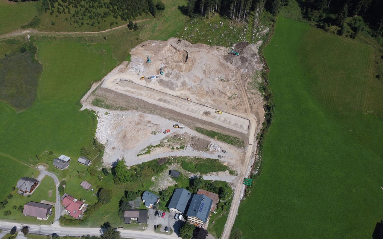 Luftaufnahme einer Baustelle in einer ländlichen Umgebung mit Häusern und grüner Wiese. Maschinen arbeiten im Vordergrund.