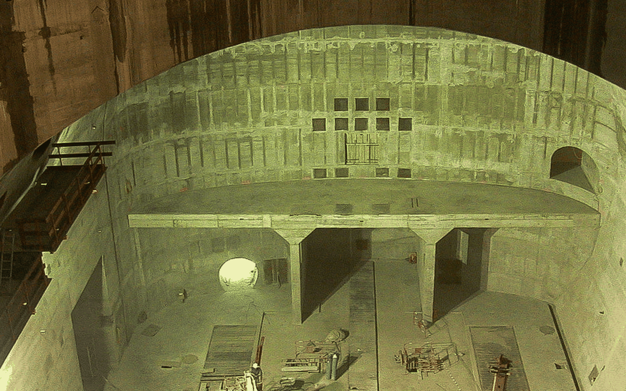 Eine große, leere, betonierte Baugrube mit Kräne und Gerüsten, die an der Wand stehen, ist zu sehen.