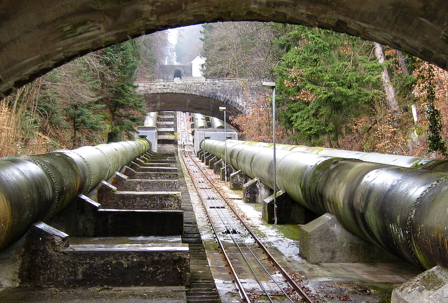Blick auf große Industrierohre, die unter mehreren Steinbogenbrücken verlaufen, umgeben von Bäumen und moosbewachsenen Strukturen.