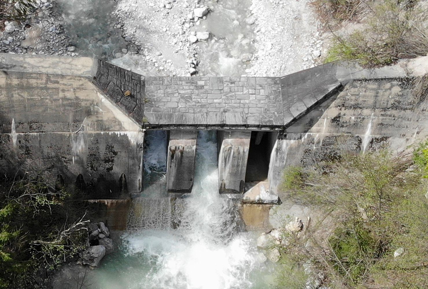 Luftaufnahme eines Betondamms mit Wasser, das durch drei Öffnungen fließt, umgeben von üppigem Grün und felsigem Gelände.