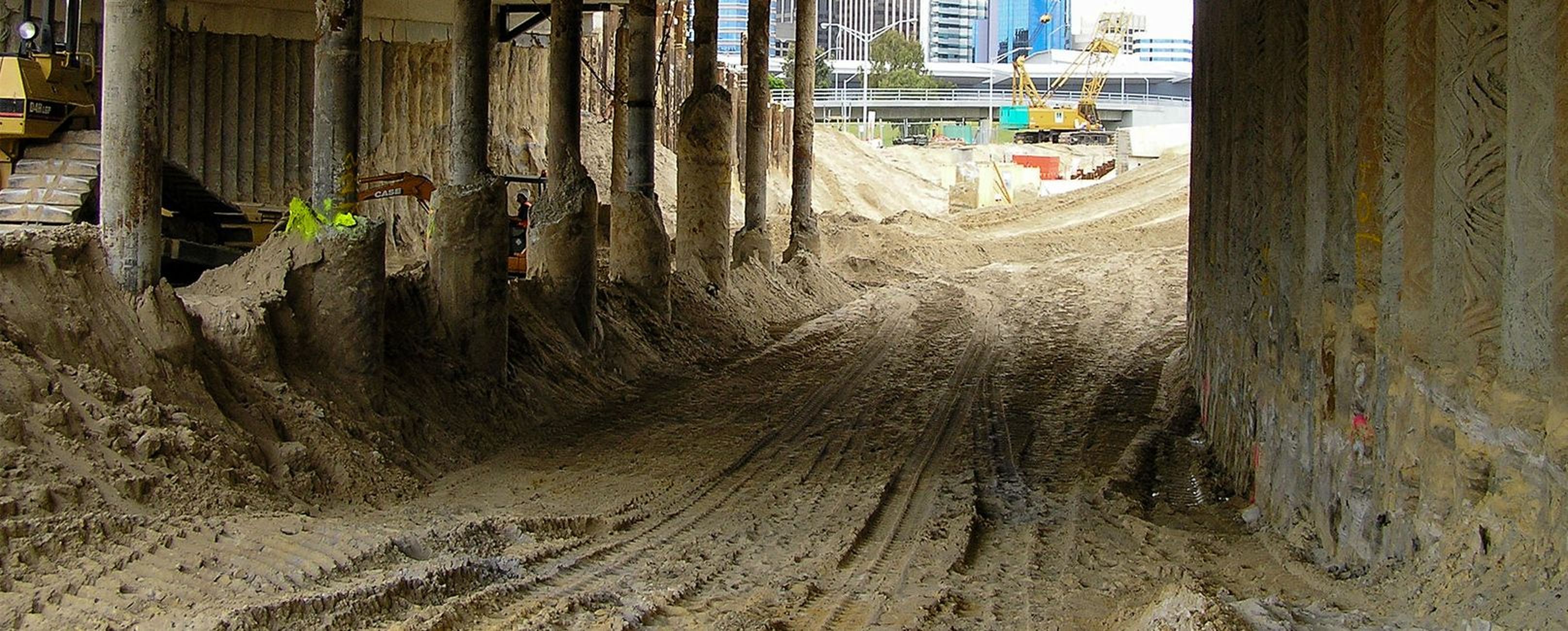 Baustelle unter einer Brücke mit sandigem Gelände, schweren Maschinen und städtischen Gebäuden im Hintergrund.