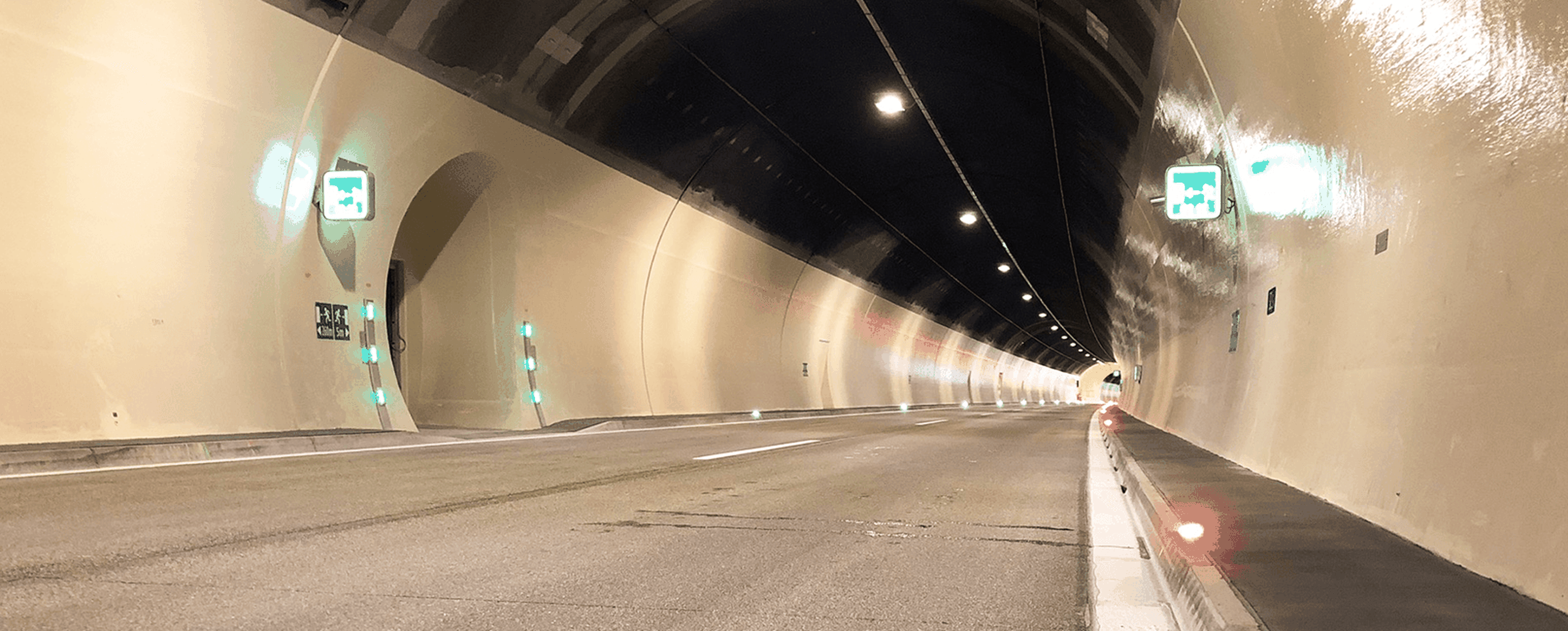 Ein gut beleuchteter, leerer Straßentunnel mit reflektierenden Wänden, grünen Ausgangsschildern und Oberlichtern, die in die Ferne reichen.