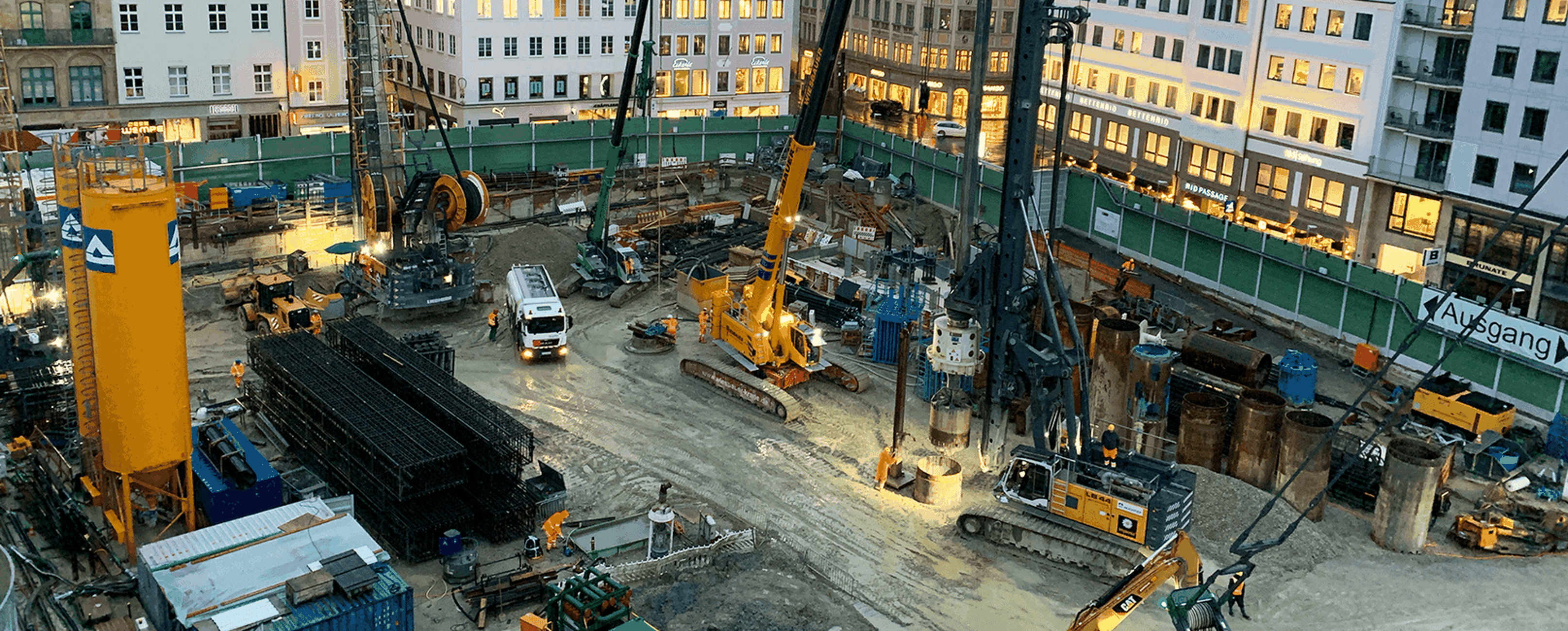 Eine Baustelle mit Maschinen und Arbeitern, umgeben von Gebäuden und einem Sicherheitszaun.