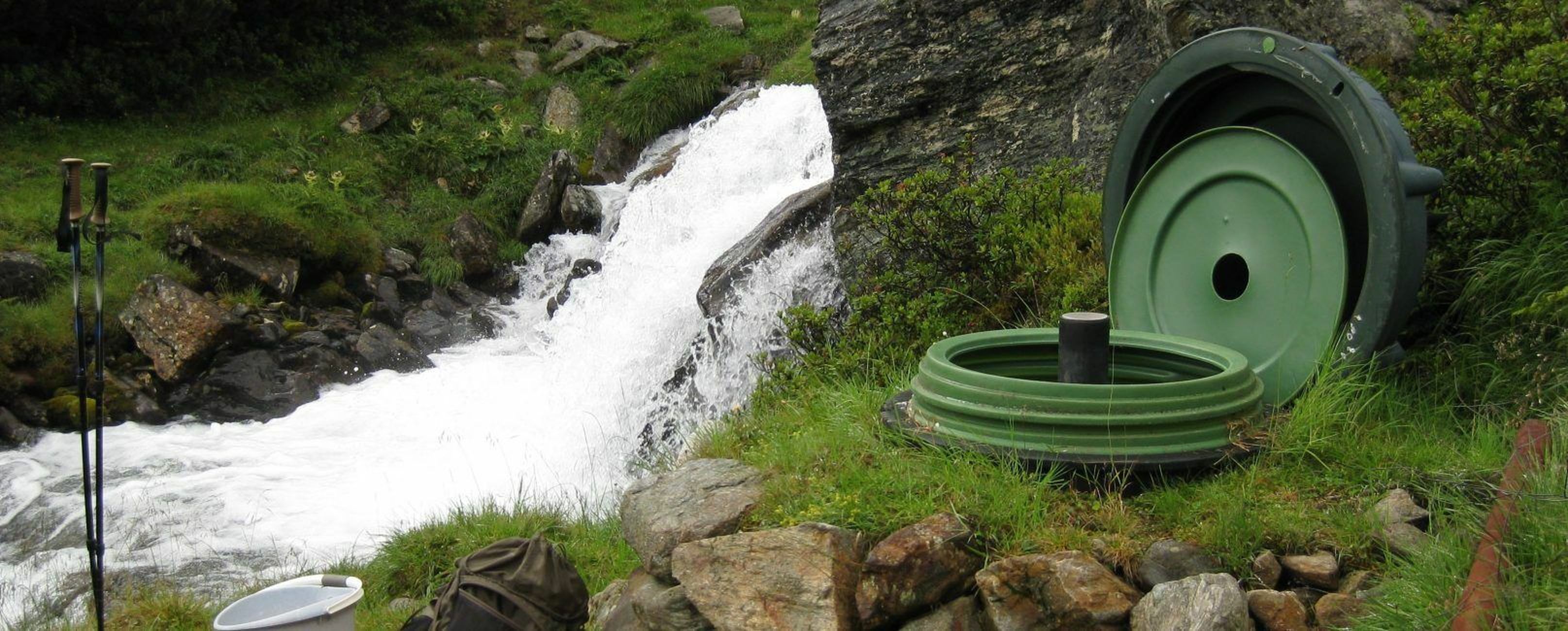 Ein kleiner Wasserfall fließt neben grünen Geräten und Felsen an einem grasbewachsenen Hang.