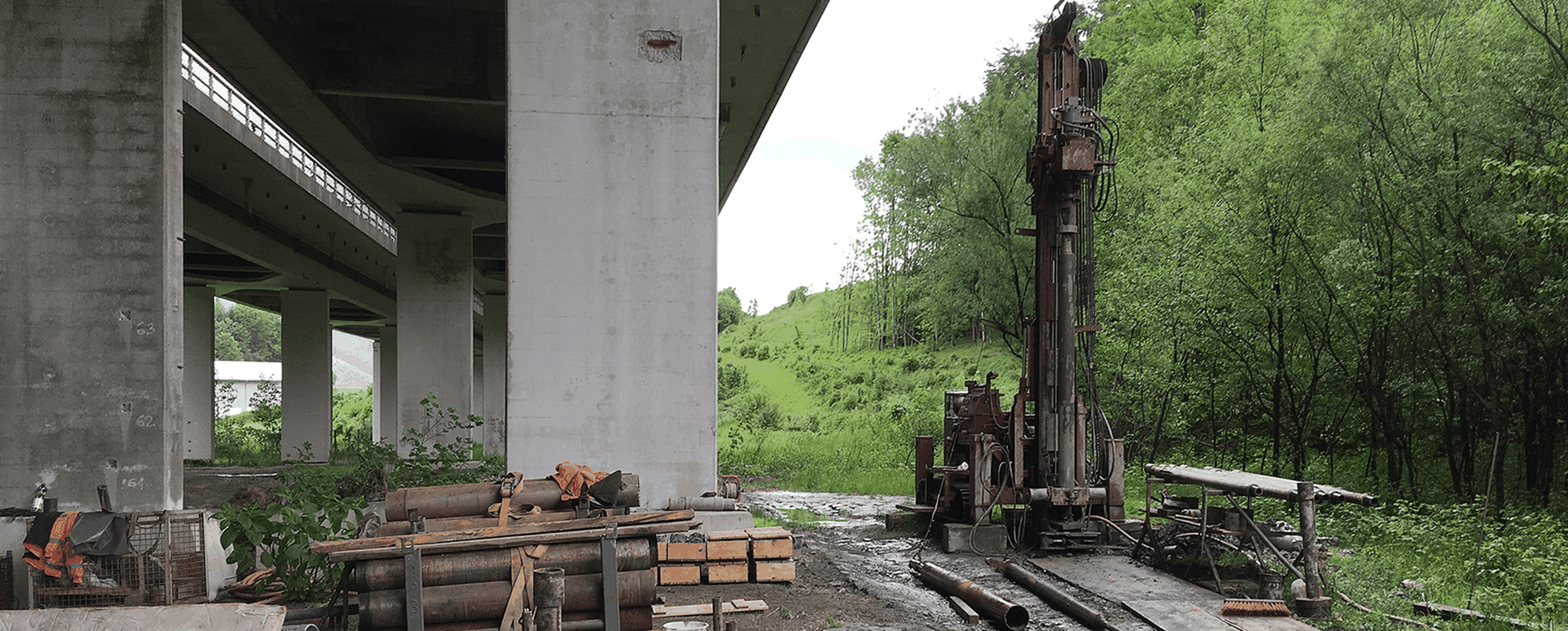 Baustelle unter einer Brücke mit Maschinen, Metallrohren und üppigen grünen Bäumen im Hintergrund.