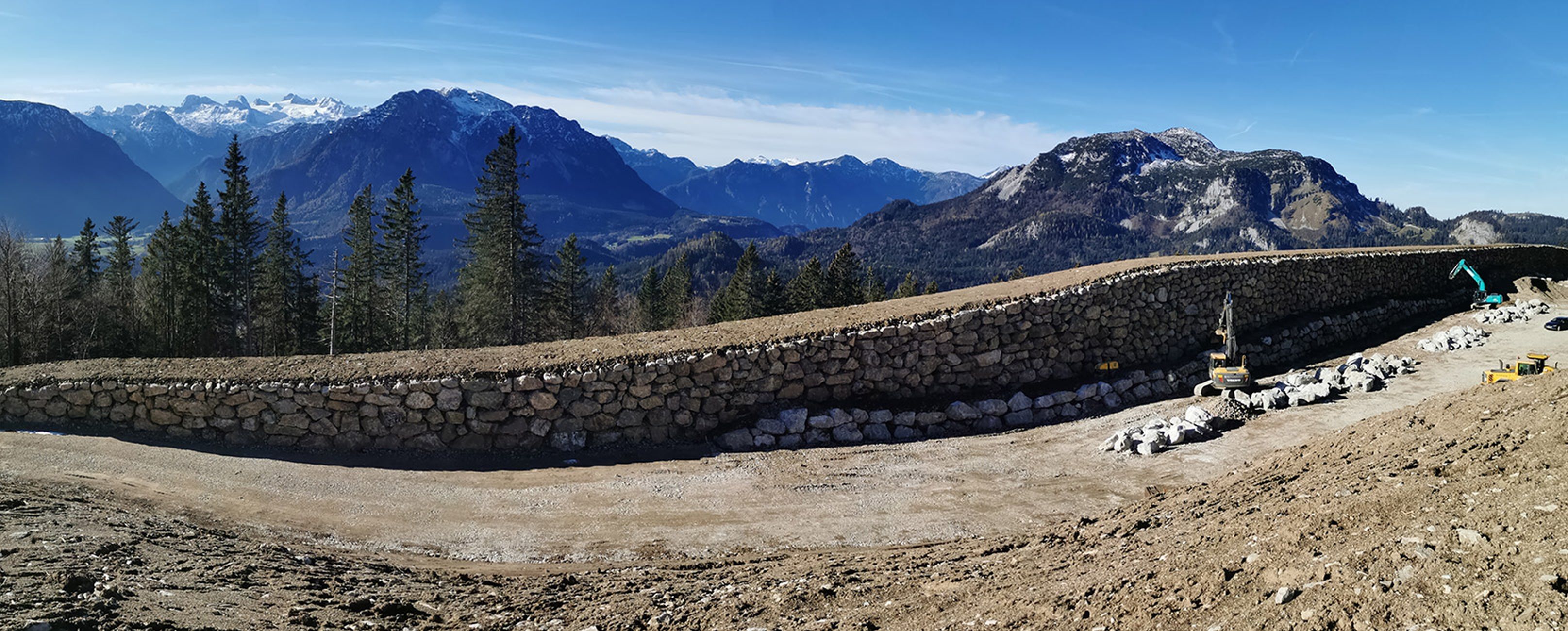 Panoramablick auf eine steinerne Mauer in den Bergen, umgeben von Bäumen, mit Baumaschinen und ohne menschliche Anwesenheit.