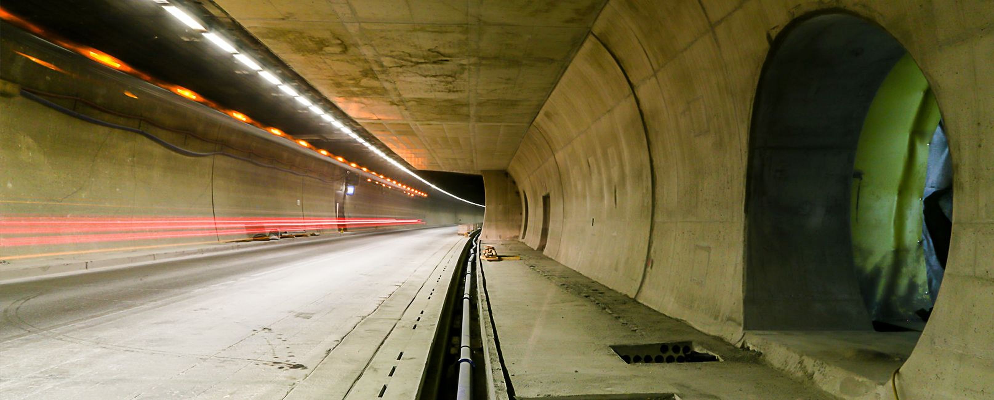 Ein Tunnel mit Betonwänden, beleuchtet von Deckenleuchten, lässt Autofahrten sichtbar werden. Rechts sind Öffnungen in der Wand.