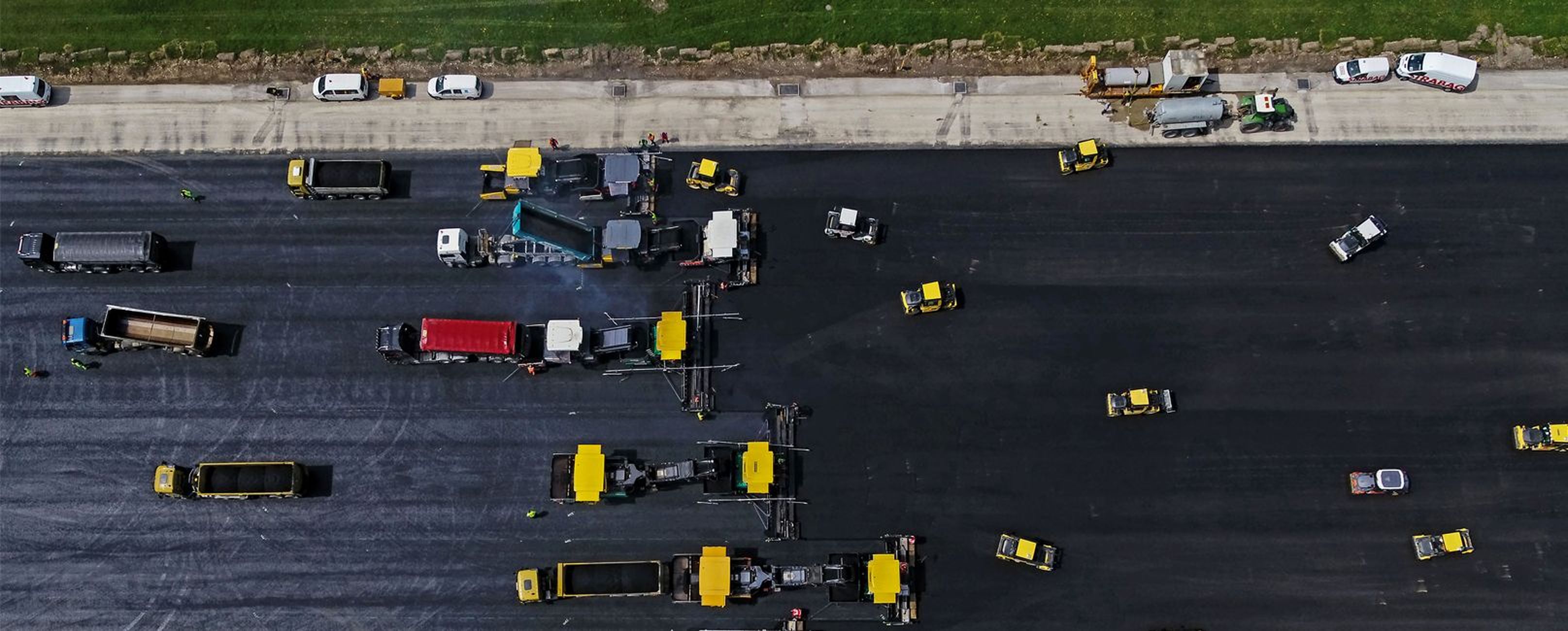 Luftaufnahme einer Straßenbaustelle mit mehreren Fahrzeugen und Geräten auf einer gepflasterten Fläche, die von Gras und geparkten Lastwagen begrenzt wird.