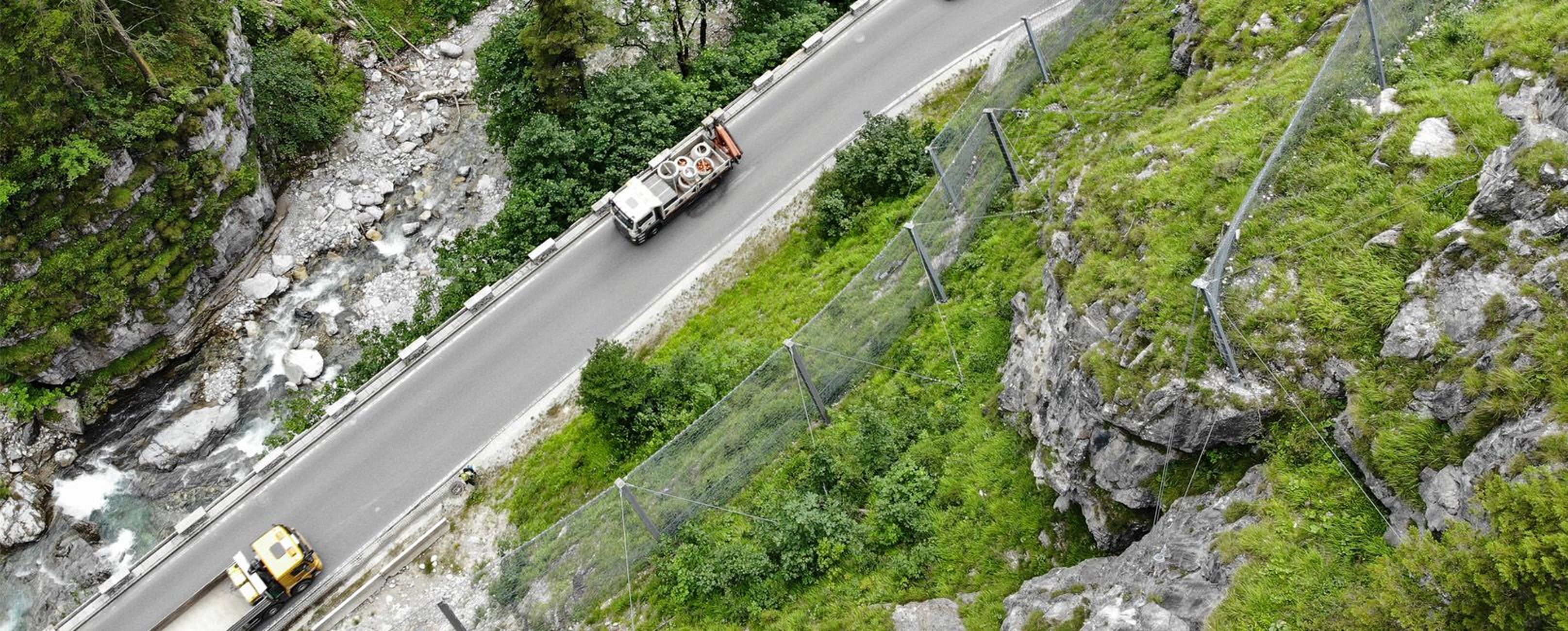 Luftaufnahme einer kurvenreichen Straße durch eine üppige, felsige Landschaft mit zwei Fahrzeugen und einem Schutzzaun am Straßenrand.