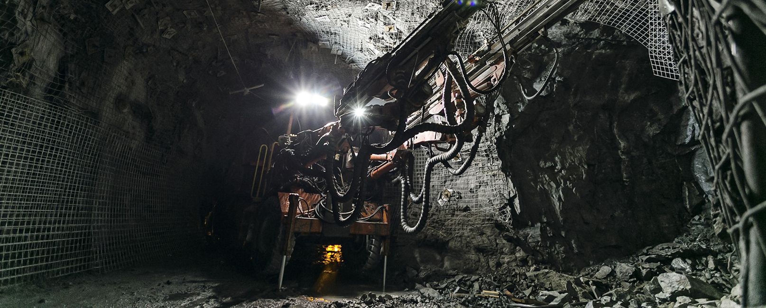 Ein Bergbaumaschinen in einem dunklen Tunnel, beleuchtet von Scheinwerfern, umgeben von felsigem Terrain.