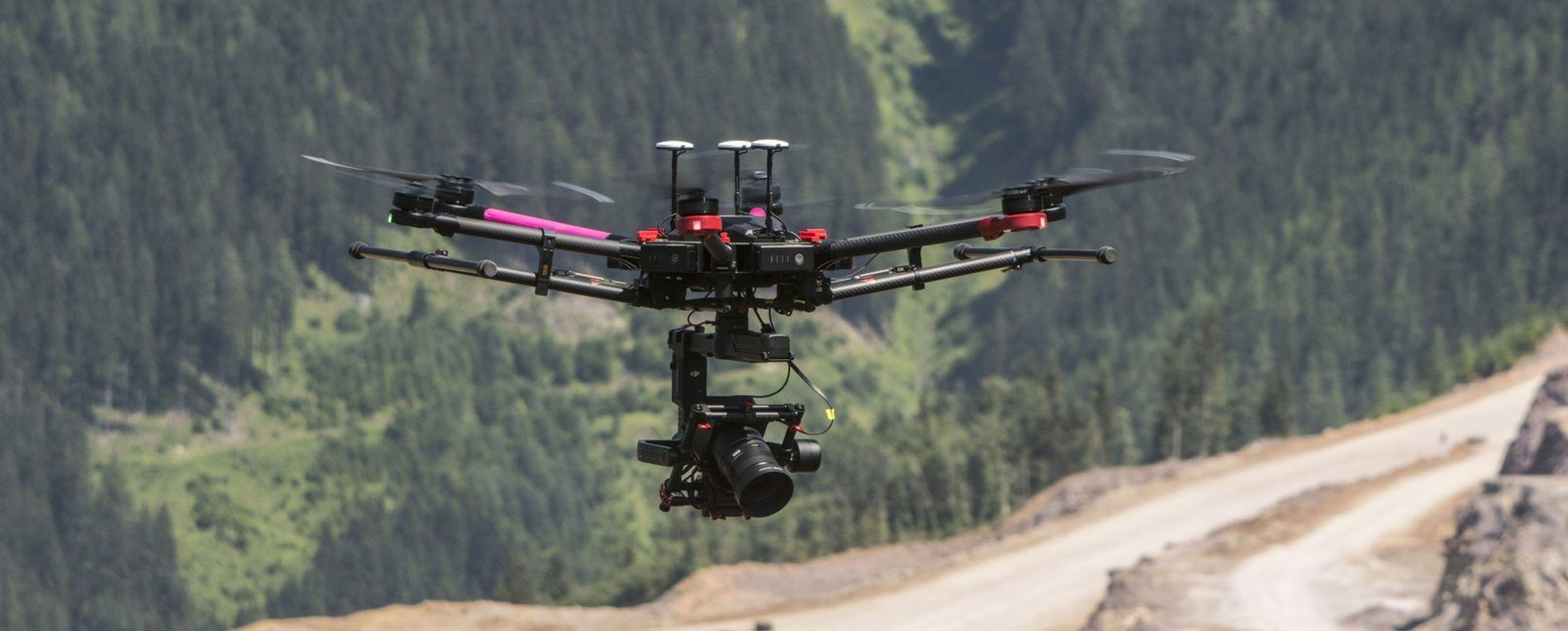 Eine mit einer Kamera ausgestattete Drohne schwebt in der Luft über einer Berglandschaft mit einer Straße und einem dichten Wald im Hintergrund.