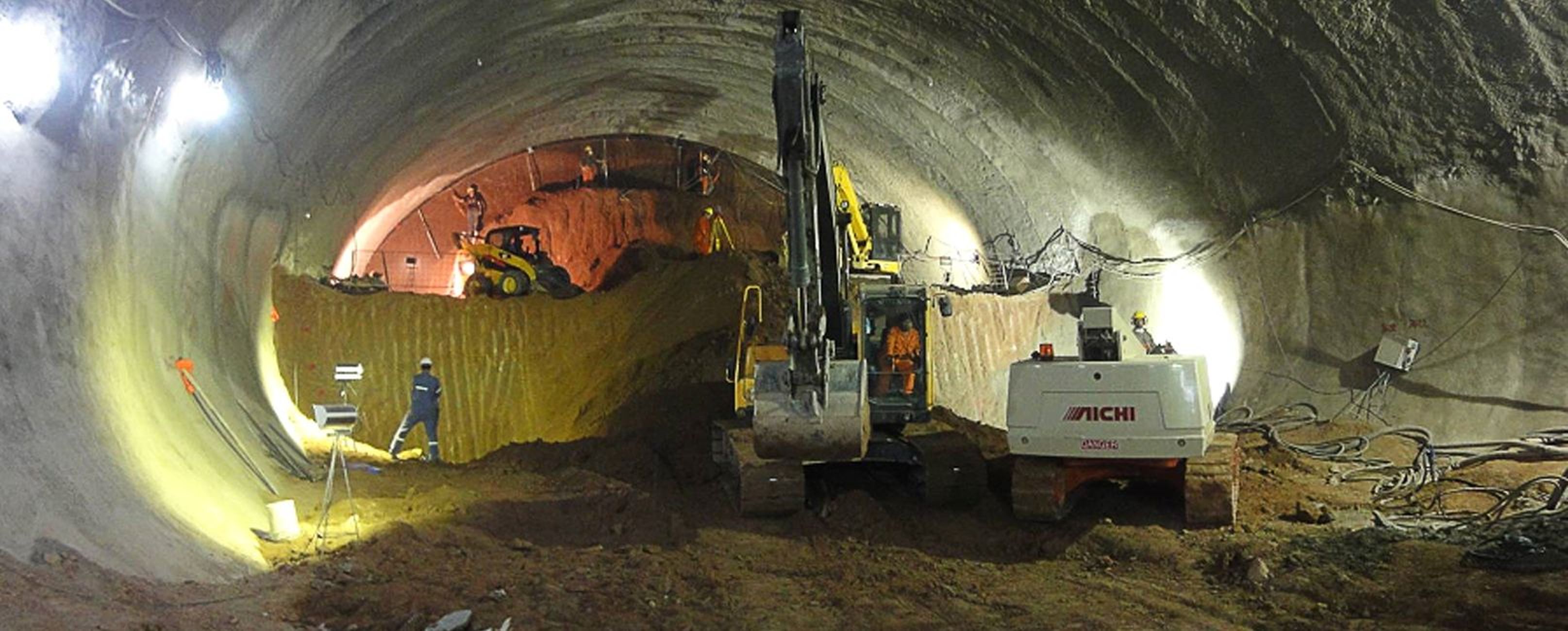Bauarbeiten in einem Tunnel mit Baumaschinen und Arbeiter*innen, beleuchtet von Scheinwerfern.
