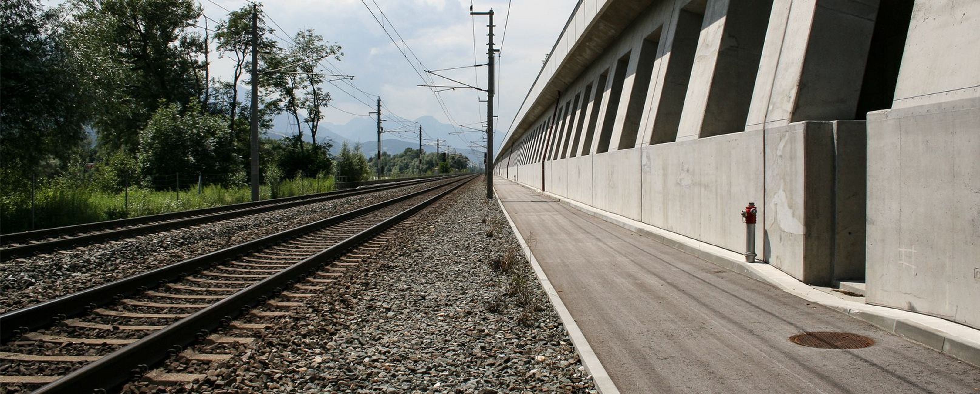 Eisenbahnschienen neben einer Betonmauer und einem Bahnsteig, mit Bäumen und Bergen im Hintergrund unter einem bewölkten Himmel.
