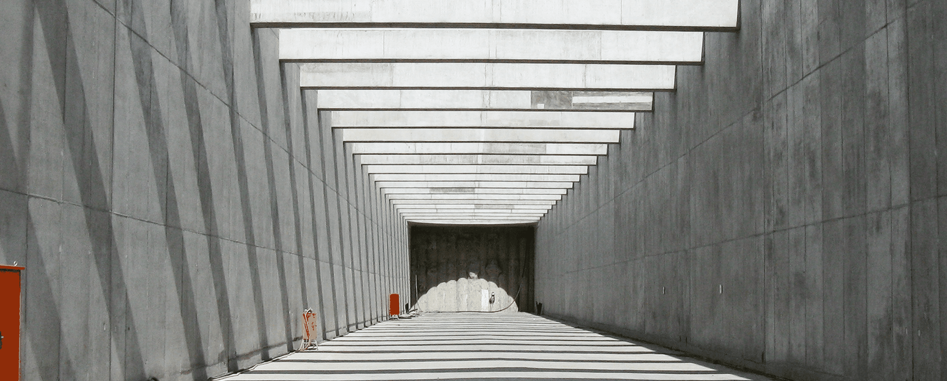 Ein langer, grauer Tunnel mit einem klaren Licht am Ende, flankiert von Betonwänden und orangefarbenen Elementen.
