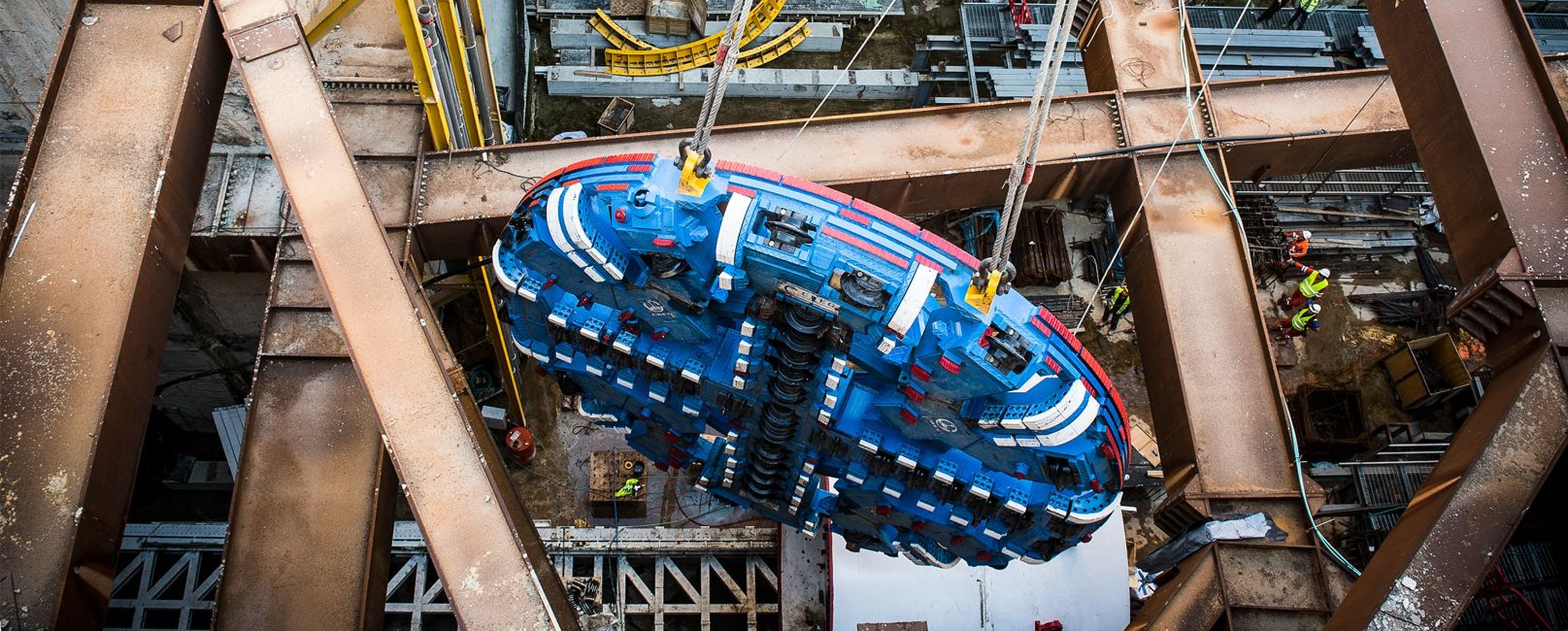 Eine große blaue Tunnelbohrmaschine wird in eine Baustelle abgesenkt, umgeben von Stahlträgern und Arbeitern in Schutzausrüstung.