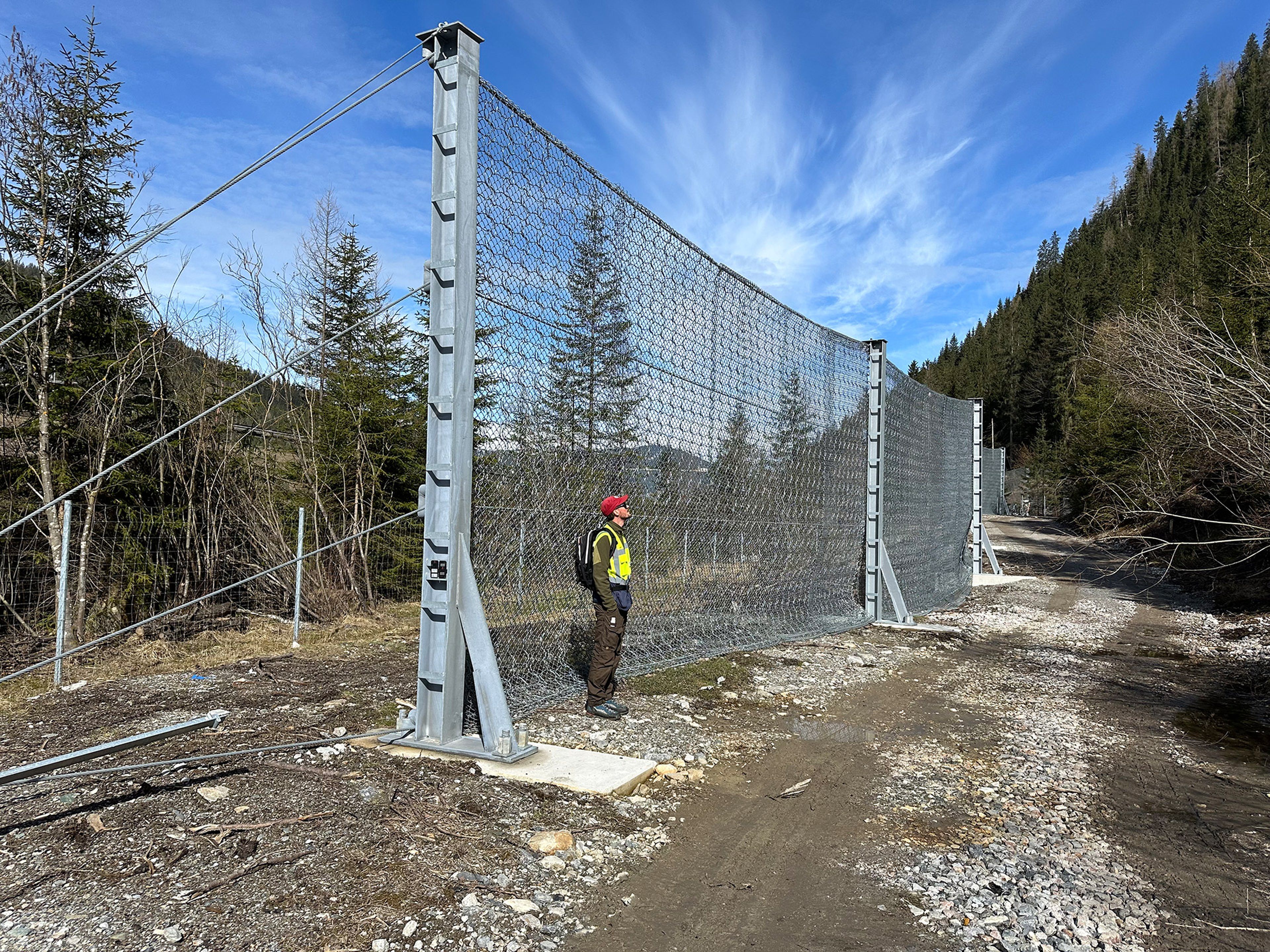 Eine Person steht an einem hohen Zaun aus Drahtgeflecht in einer ländlichen Umgebung mit Bäumen und einem blauen Himmel.
