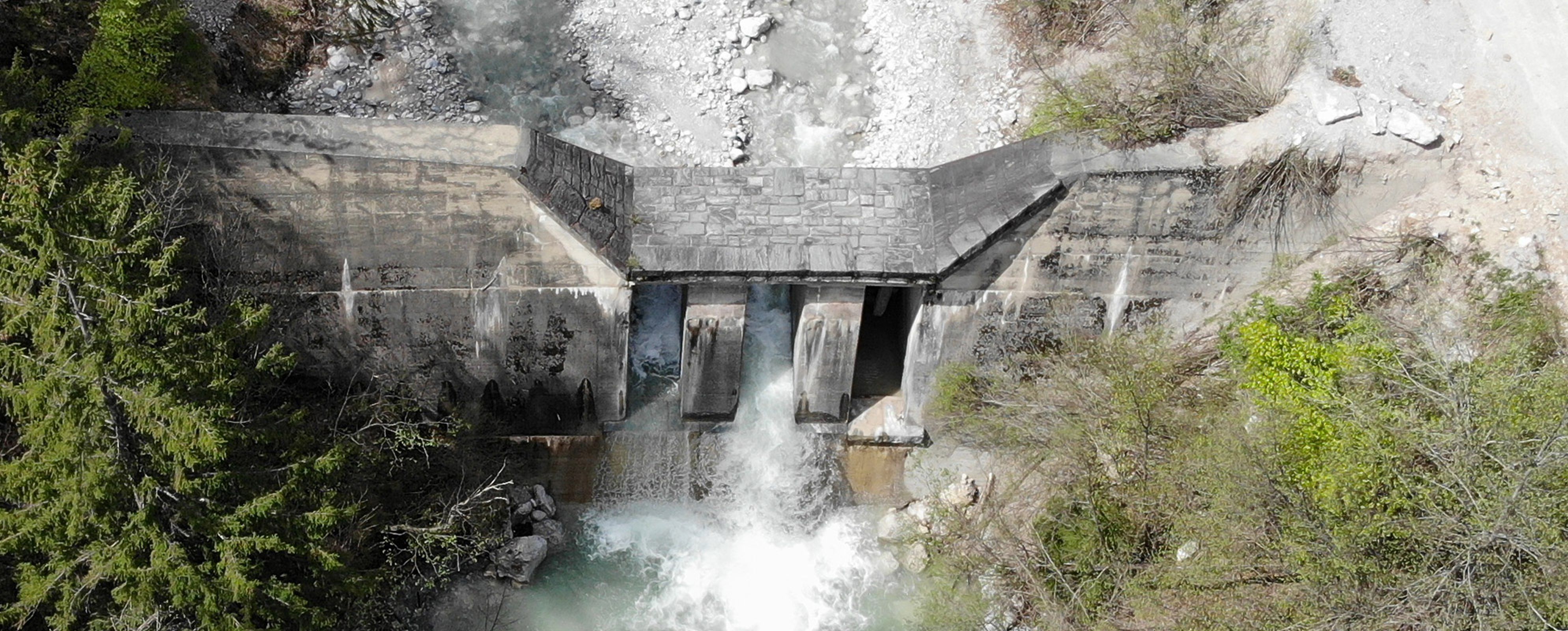 Aerial view of a concrete dam with water flowing through three openings, surrounded by lush greenery and rocky terrain.