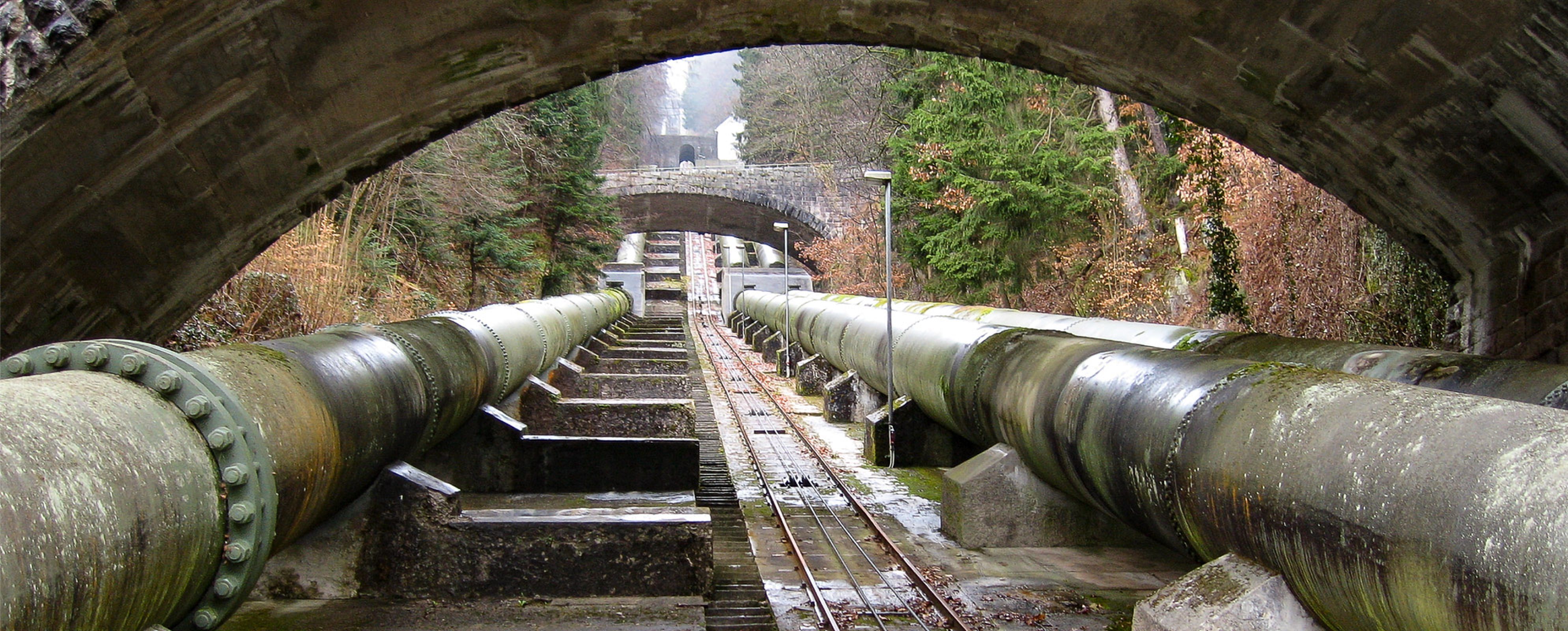 View of large industrial pipes running under several stone arch bridges, surrounded by trees and moss-covered structures.