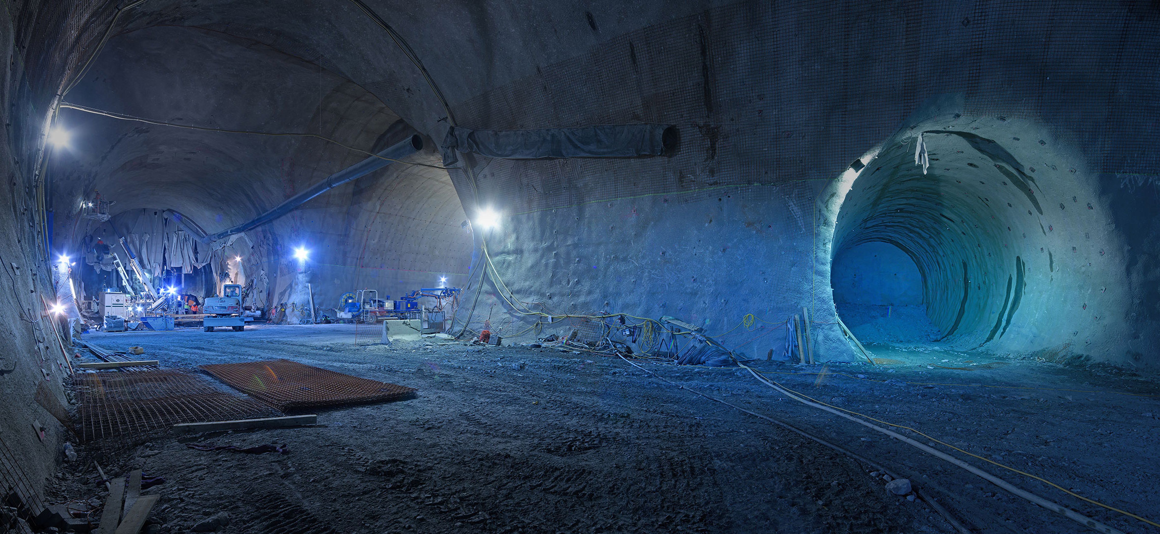 Ein großer Tunnel mit Baustellenfahrzeugen und Beleuchtung. Der Boden ist uneben, und im Hintergrund sieht man einen Tunnelausgang.