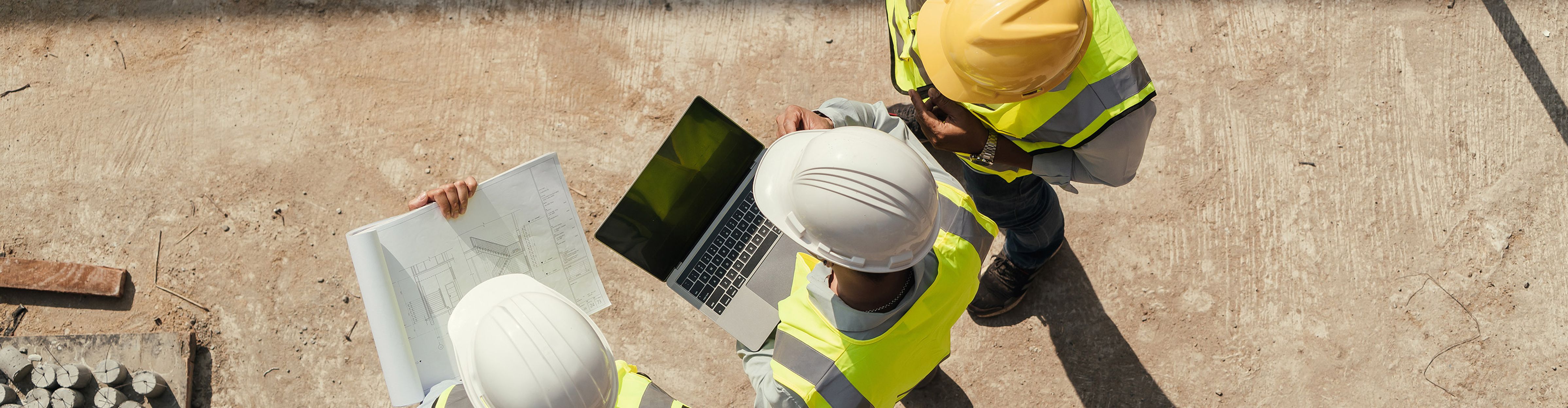 Drei Bauarbeiter in Schutzkleidung prüfen Pläne und einen Laptop auf einer Baustelle.
