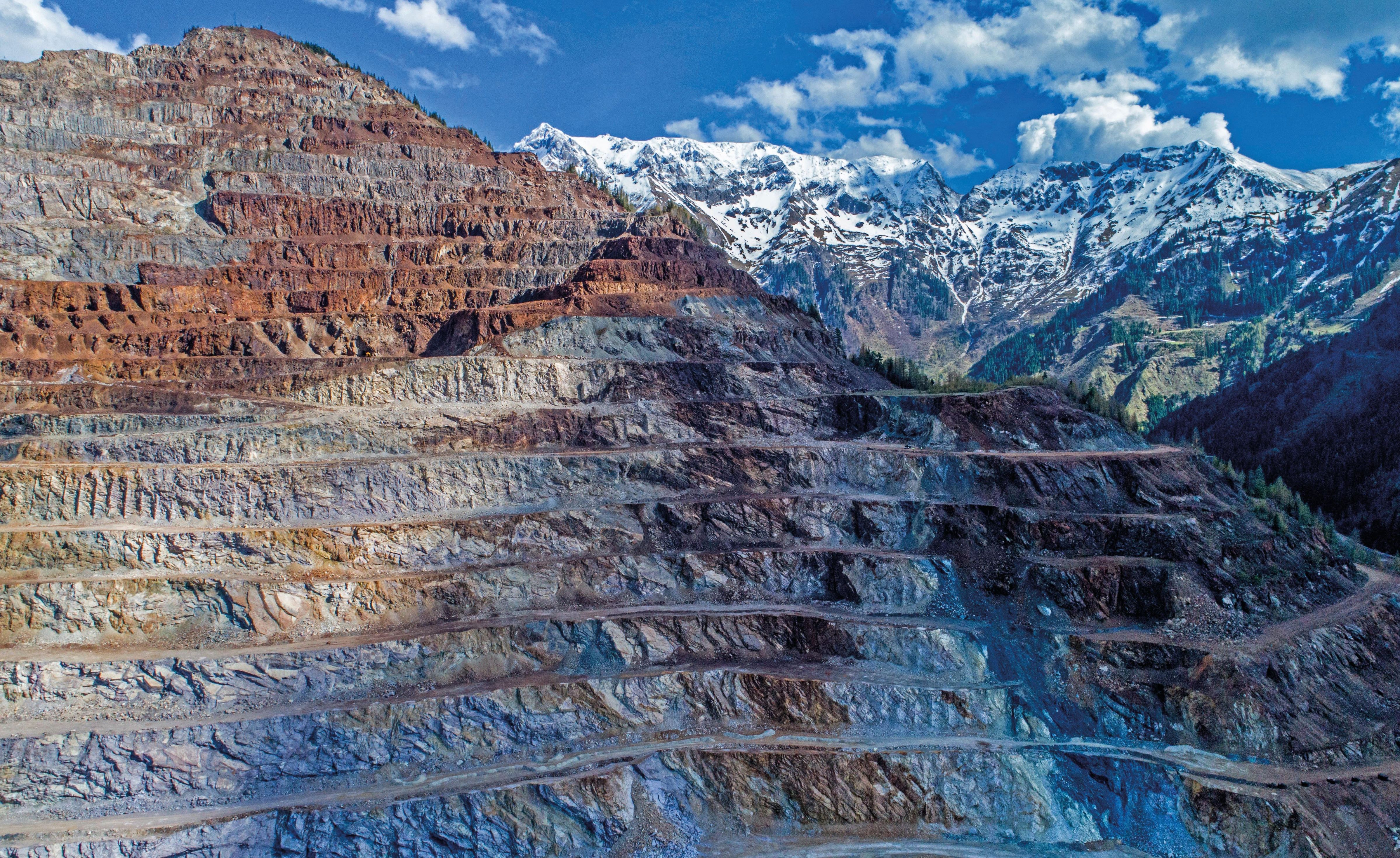 Terrassenförmiger Tagebau mit geschichteten Felsformationen vor dem Hintergrund schneebedeckter Berge und eines teilweise bewölkten Himmels.