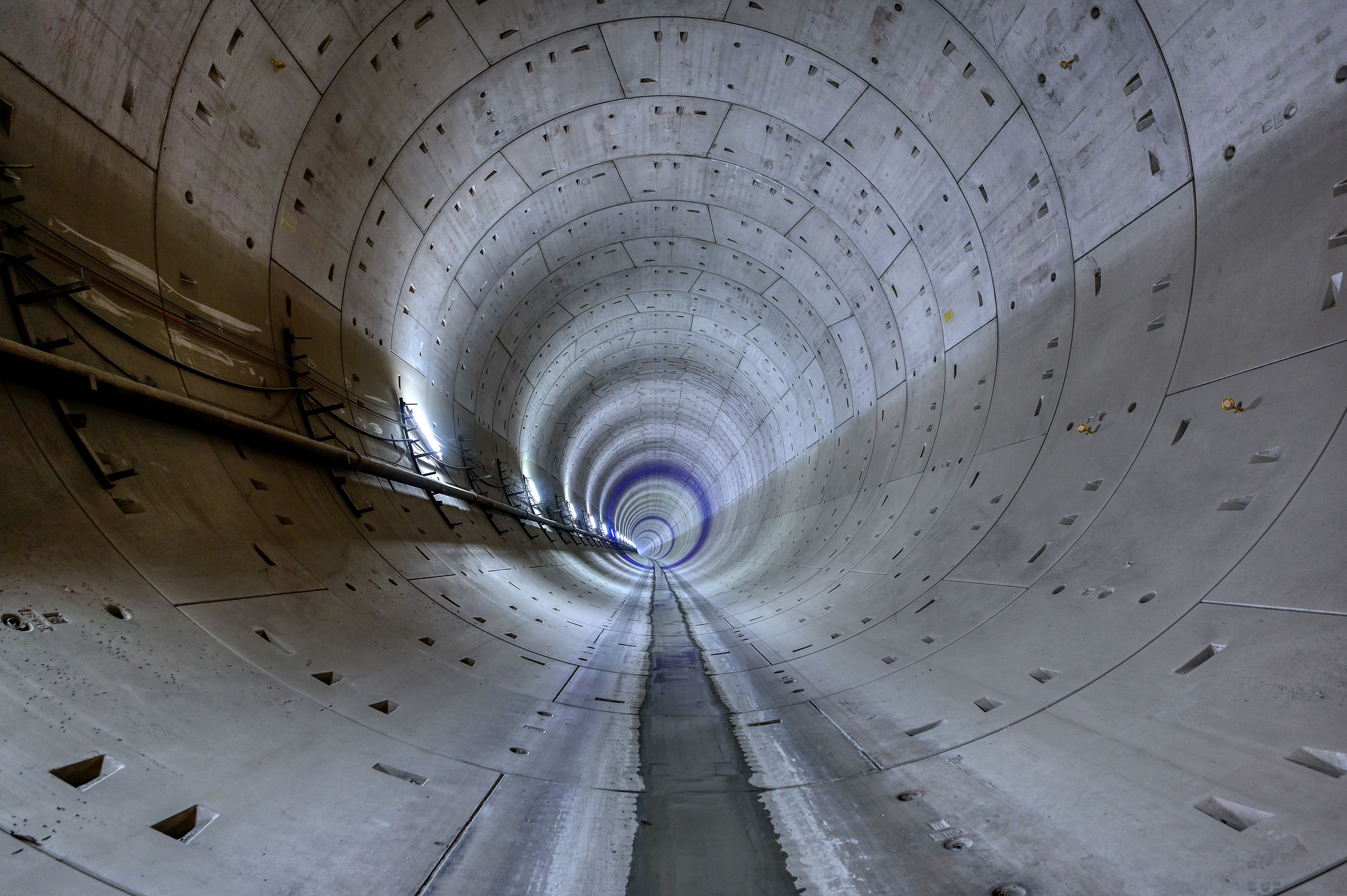 Innenansicht eines großen, kreisförmigen Betontunnels mit glattem Boden, der sich in die Ferne erstreckt und an der linken Wand von Kabeln gesäumt ist.