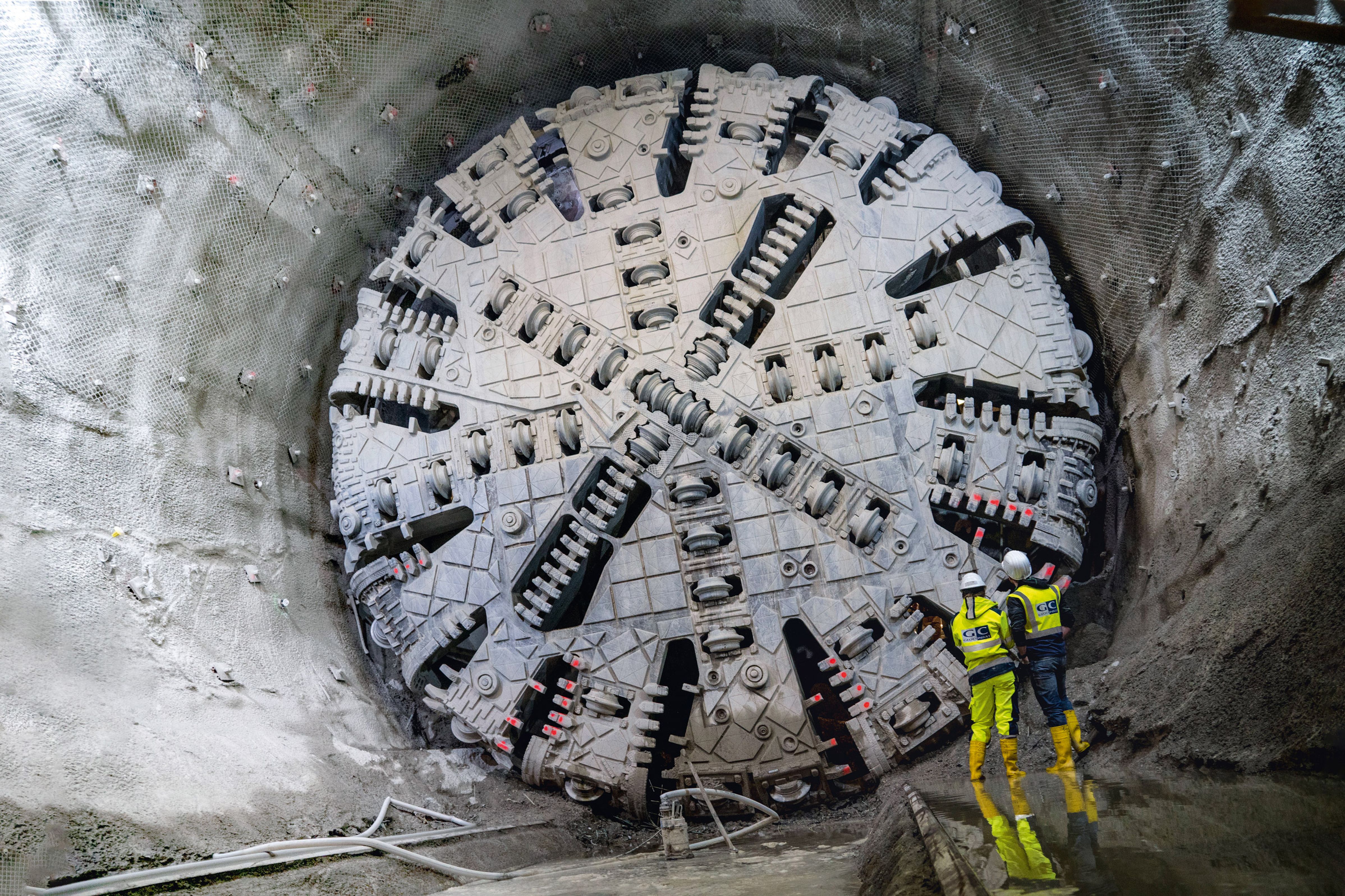 Zwei Arbeiter in Schutzausrüstung stehen vor einer großen Tunnelbohrmaschine in einem teilweise ausgebrochenen Tunnel.