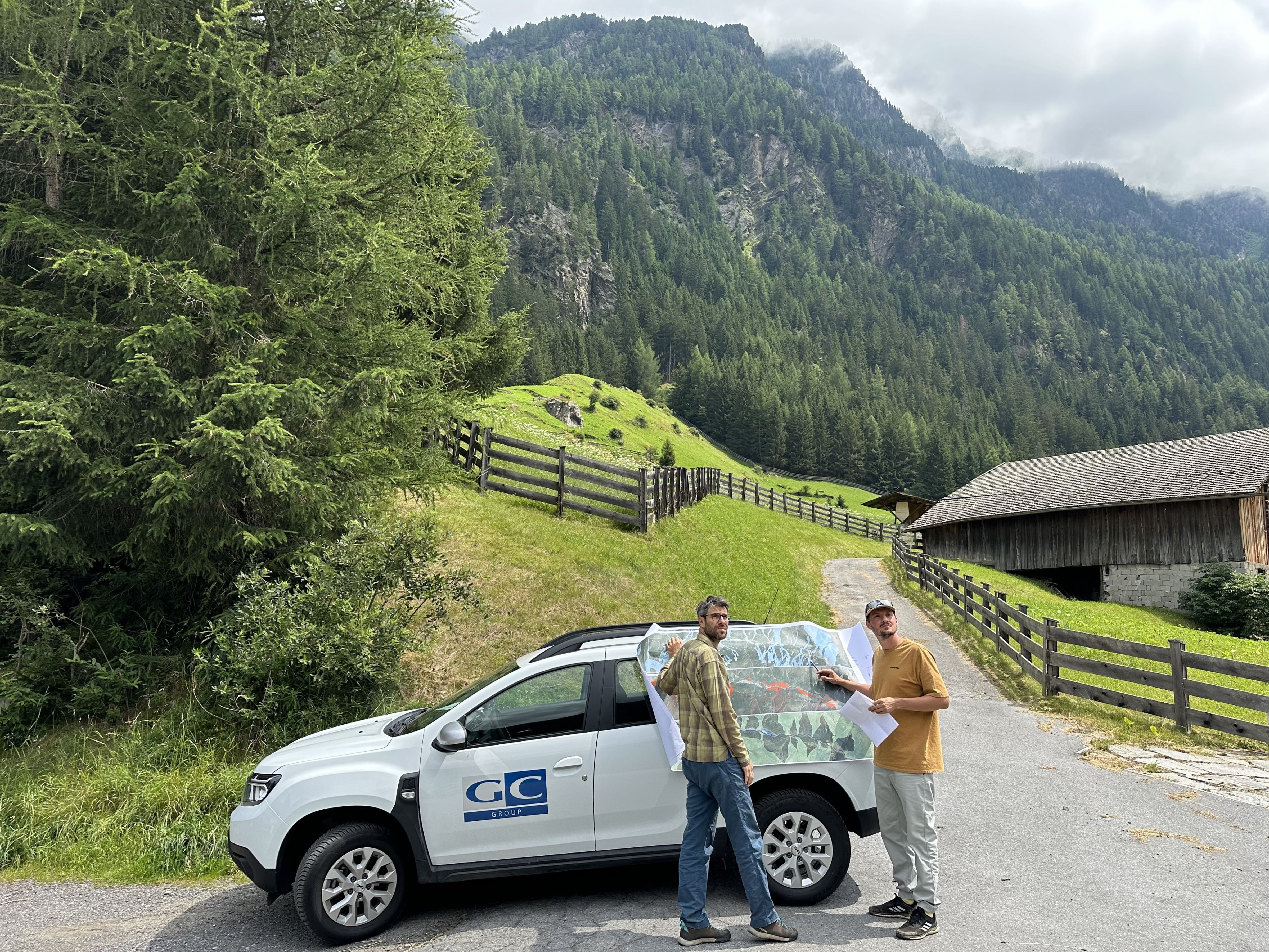 Zwei Personen betrachten eine Karte an einem Auto in einer ländlichen Umgebung mit Bergen und Wäldern.
