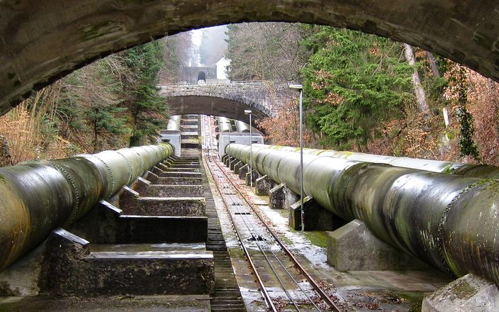 Blick auf große Industrierohre, die unter mehreren Steinbogenbrücken verlaufen, umgeben von Bäumen und moosbewachsenen Strukturen.