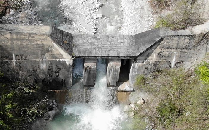 Luftaufnahme eines Betondamms mit Wasser, das durch drei Öffnungen fließt, umgeben von üppigem Grün und felsigem Gelände.