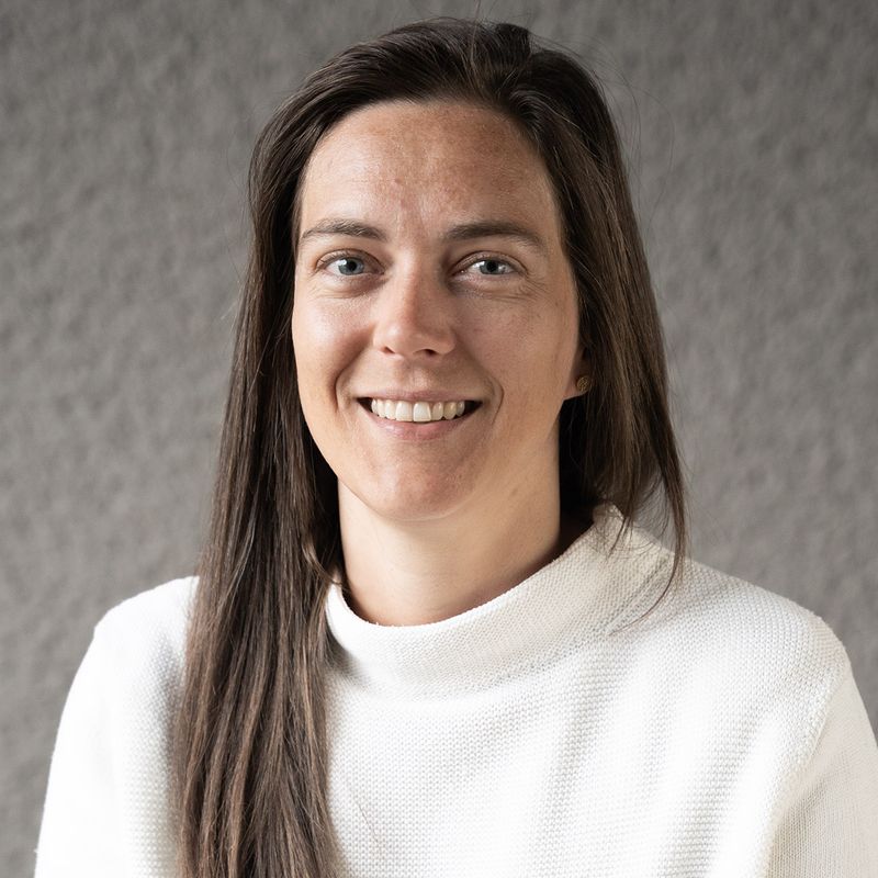 Lächelnde Frau mit langen braunen Haaren, die einen weißen Pullover trägt und vor einem strukturierten grauen Hintergrund steht.