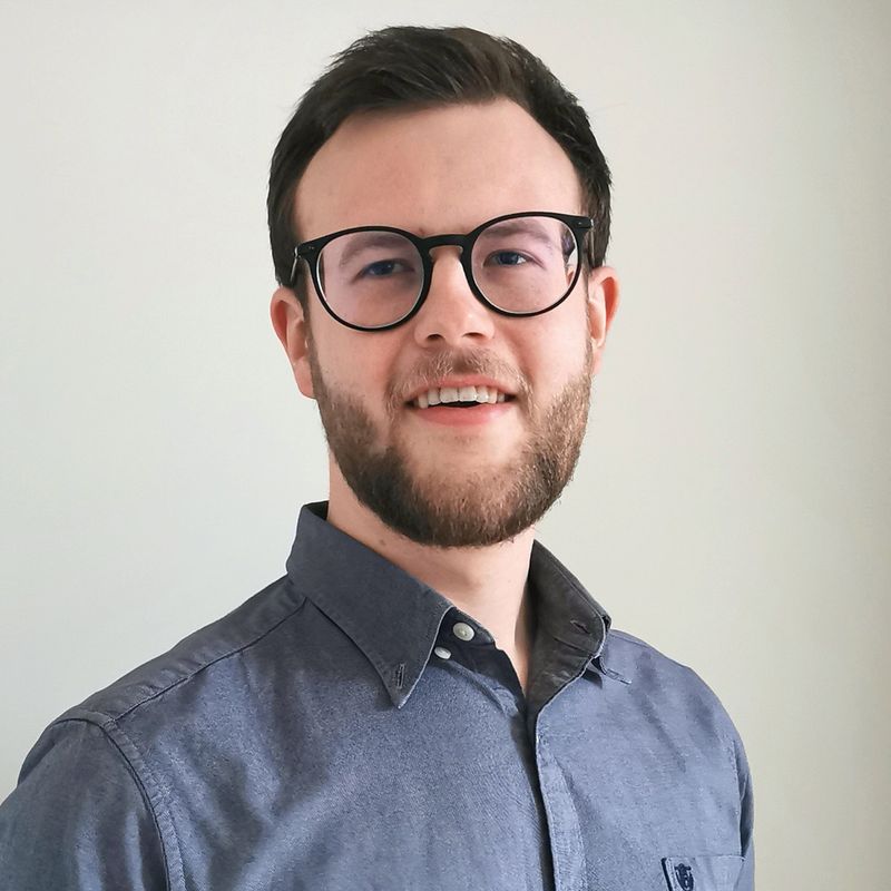 Ein Mann mit kurzen Haaren, einer Brille und einem blauen Hemd mit Knöpfen lächelt vor einem schlichten Hintergrund.